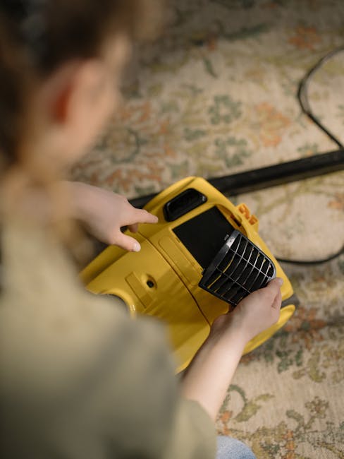 Close-up image showing a person operating a yellow portable industrial fan for surface cleaning or drying purposes on a patterned carpet in a room. The person's hands are on the fan, with their face blurred in the foreground. The carpet displays a floral pattern in muted tones, and the room is illuminated with natural light, highlighting the clean, dry condition of the surface. Visible in the background are electrical cords connected to the fan, indicating ongoing deep cleaning or sanitisation work. The scene reflects professional cleaning services provided by Carpet Cleaning Hammersmith, focusing on maintaining hygiene and cleanliness during domestic or commercial cleaning tasks.