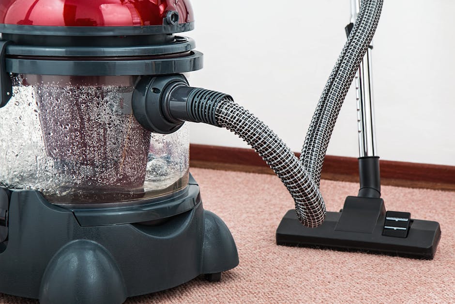 Close-up of a modern vacuum cleaner with a red and black body, clear dust container, and a flexible hose connected to a flat, rectangular cleaning head resting on a light pink carpet in a room with white walls and wooden baseboards, demonstrating surface cleaning and deep cleaning processes used by Carpet Cleaning Hammersmith for domestic or commercial hygiene maintenance.