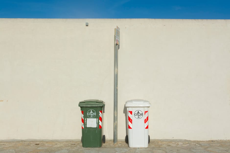 Two cylindrical waste bins mounted on metal frames are positioned side-by-side on a beige concrete surface. The bin on the left is bright yellow with small perforated holes, designated for general was