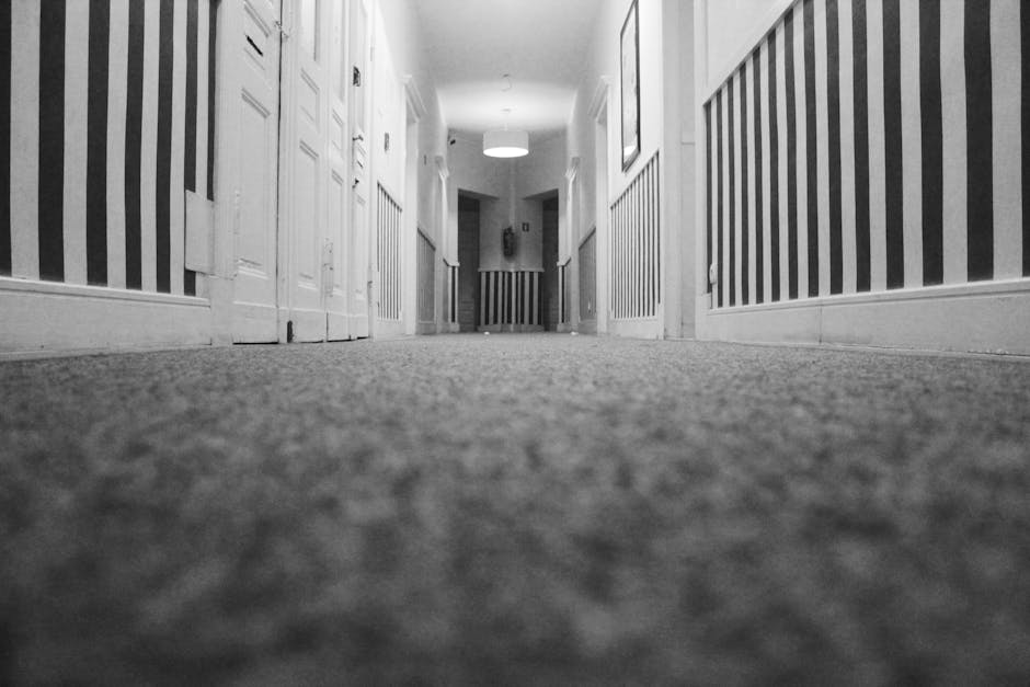 A black and white photo of a corridor inside a residential flat, taken from a low angle close to the carpeted floor. The corridor features striped wallpaper on the walls, with a white wooden dado rail