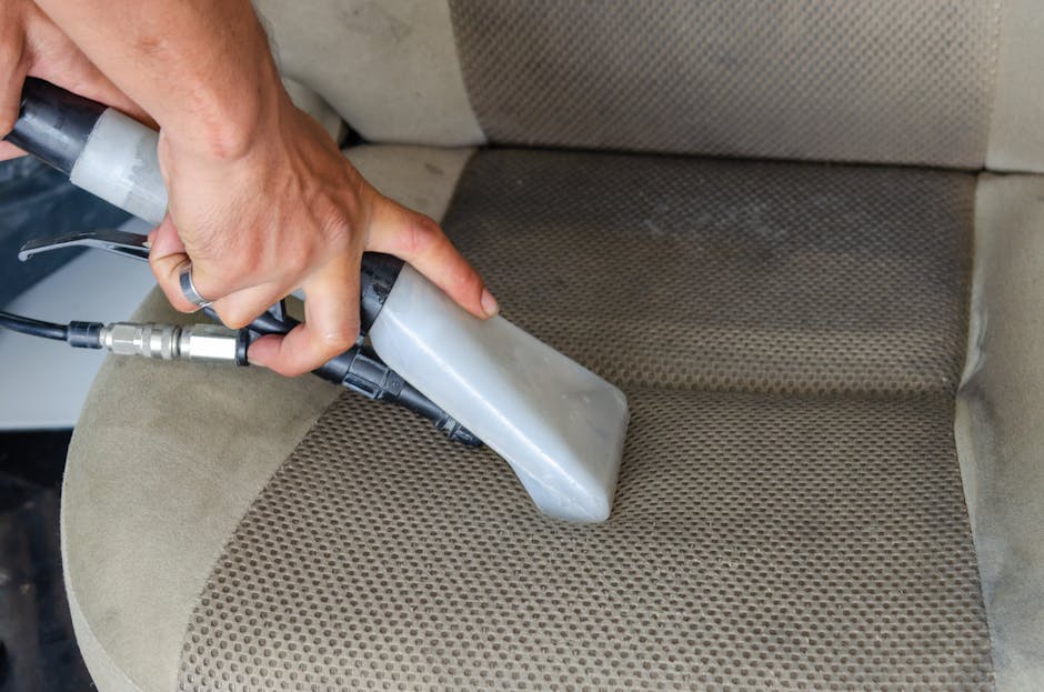 A close-up of a hand holding a steam cleaning tool positioned over the fabric upholstery of a sofa in a residential setting. The upholstery features a tan, patterned cloth material with visible dirt and stains, especially on the seat cushion. The steam cleaner, connected to a black and grey hose, is being used to deep clean the fabric surface, with the surrounding area appearing well-lit and clean. The scene is indicative of surface cleaning and deep cleaning processes performed by Carpet Cleaning Hammersmith, reflecting their expertise in domestic cleaning and upholstery sanitisation.
